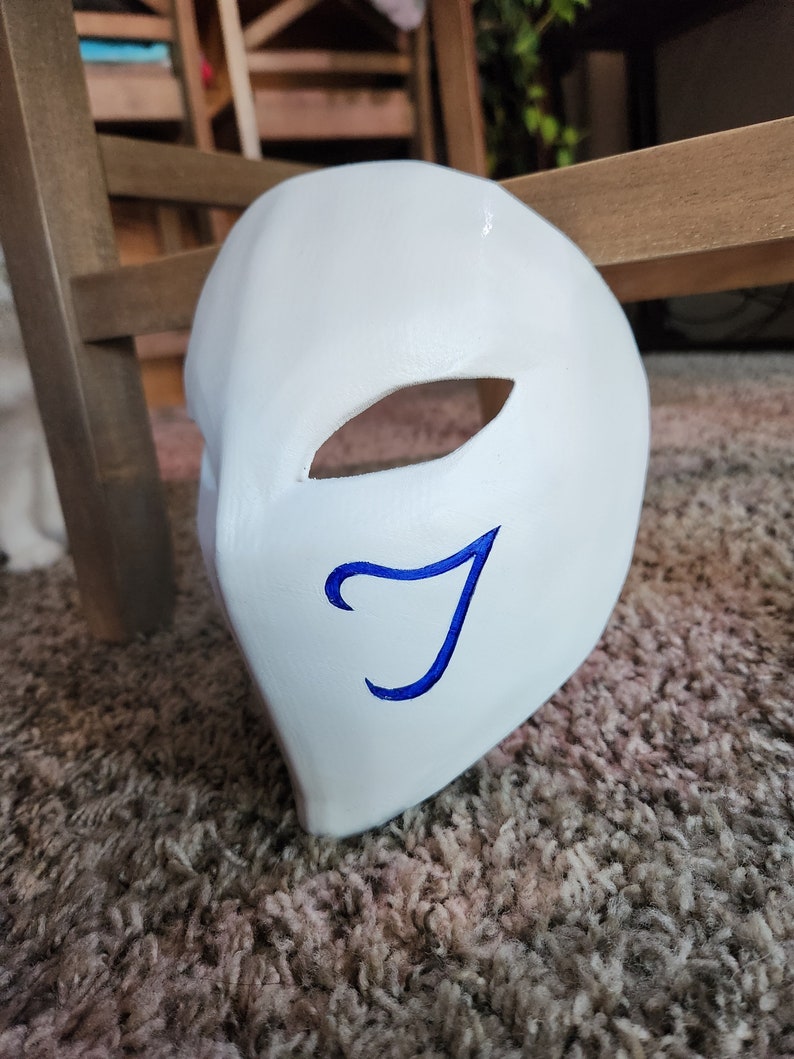 May include: A white mask with a blue letter 'J' painted on it. The mask has a single eye hole.