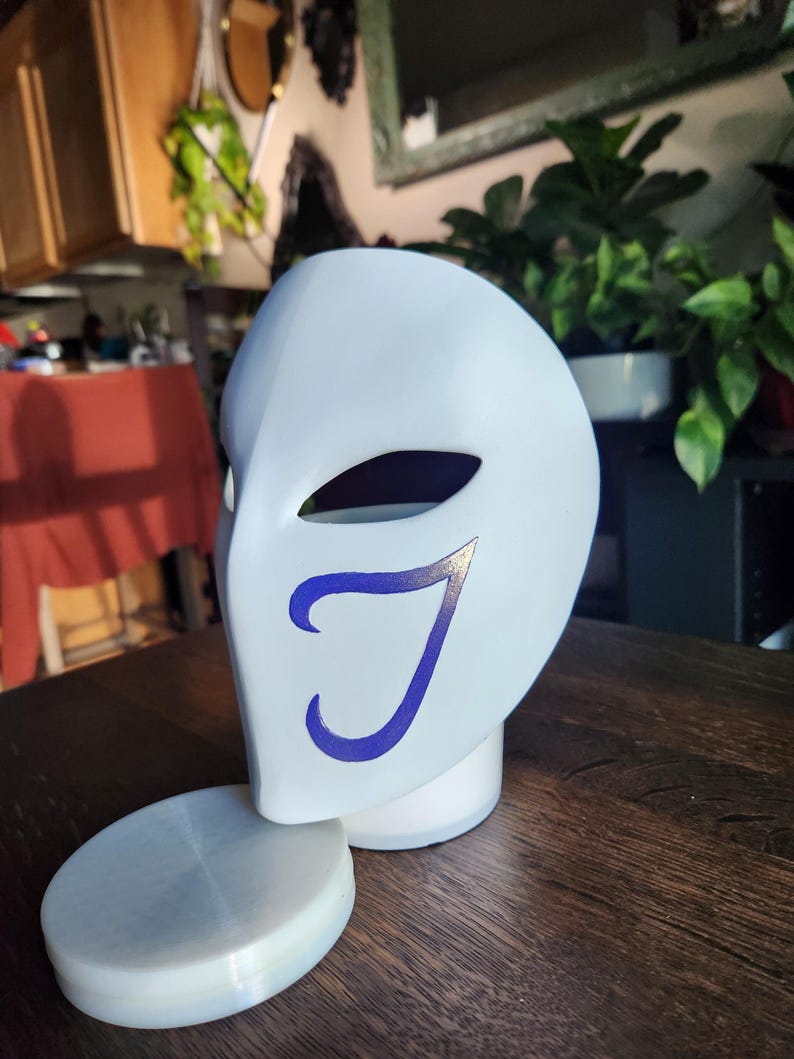 May include: A white mask with a blue 'J' design. The mask is sitting on a white base.