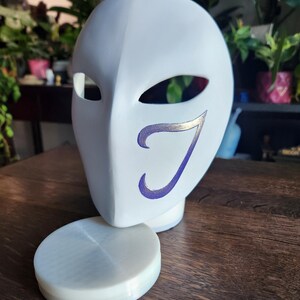 May include: White plastic mask with a purple and gold design. The mask is sitting on a white plastic stand.