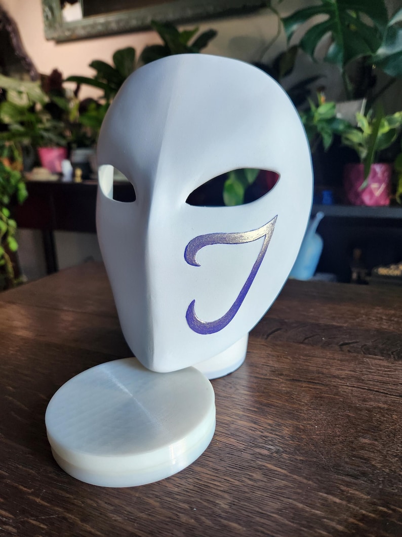 May include: White plastic mask with a purple and gold design. The mask is sitting on a white plastic stand.