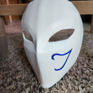 May include: White mask with a blue swirl design. The mask has two eye holes and a curved shape.