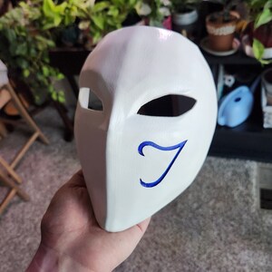 May include: White mask with a blue stylized letter J on the right side. The mask has two eye holes and a pointed chin.