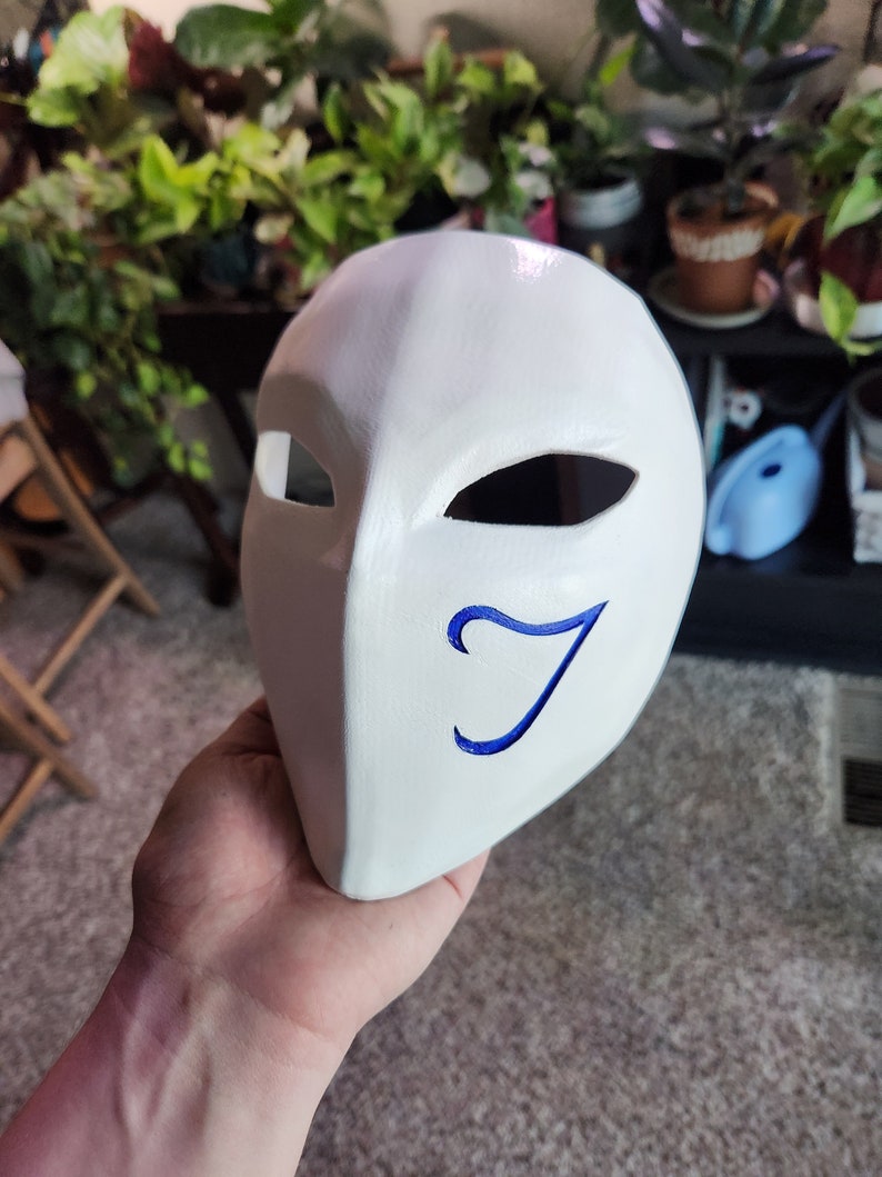 May include: White mask with a blue stylized letter J on the right side. The mask has two eye holes and a pointed chin.