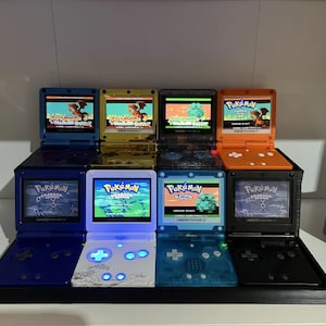 May include: A collection of eight different colored Game Boy Advance SP consoles, each with a different Pokemon game cartridge inserted. The consoles are arranged in a row on a black display stand. The games include Pokemon Emerald, Pokemon Diamond, Pokemon Platinum, and Pokemon Ruby.