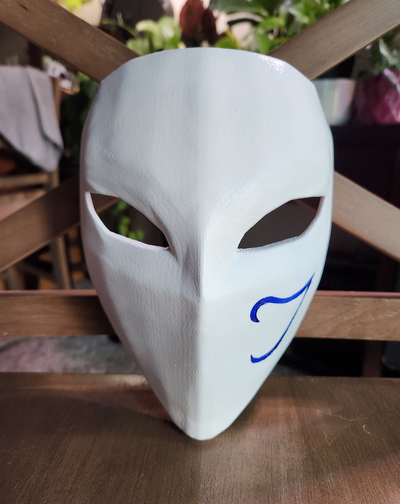 May include: White mask with a blue swirl design on the right side. The mask has two eye holes and a pointed chin.