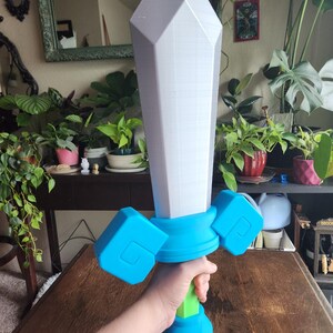 May include: A large, white, plastic sword with a blue and green handle. The sword is held by a hand in the image.