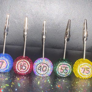 May include: Five bedazzled billiard ball-themed photo holders. Each holder features a different number: 7, 16, 40, 55, and 75. The balls are in blue, red, purple, green, and yellow, with silver clips.