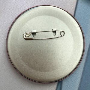 May include: A circular metal pin-back button featuring a smooth, silver-toned finish on the reverse side. A horizontal steel safety pin mechanism is securely attached across the center, designed for fastening the button to fabric or clothing.