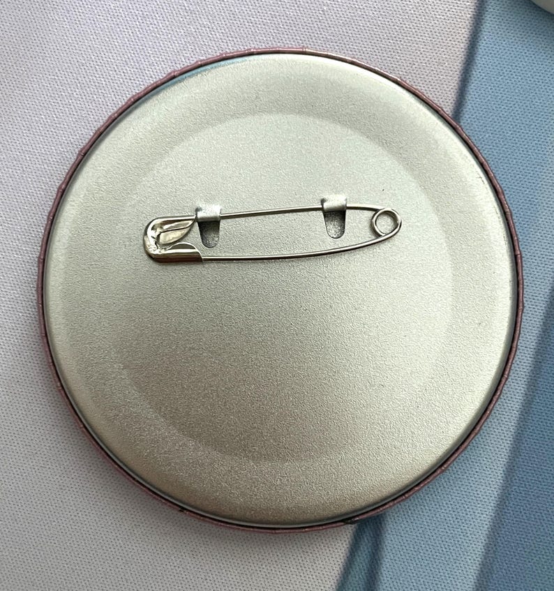 May include: A circular metal pin-back button featuring a smooth, silver-toned finish on the reverse side. A horizontal steel safety pin mechanism is securely attached across the center, designed for fastening the button to fabric or clothing.