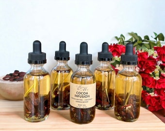 May include: Five small glass bottles with black dropper tops filled with a golden liquid and containing brown pieces of fruit. The bottles are labeled "Cocoa Infusion Cocoa Butter - Coconut - Sandalwood Body Oil 1 fl. oz. / 30 ml."