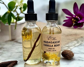 Op de afbeelding: Twee doorzichtige glazen flessen met zwarte druppelaars, gevuld met een lichtgele vloeistof. De flessen bevatten gedroogde bloemen en een vanilleboon. Het etiket op de fles staat "Madagascar Vanilla Bean Body Oil 2 fl. oz. | 60 ml."