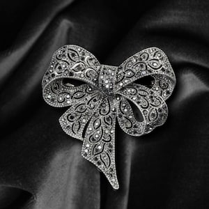 Op de afbeelding: Een zilverkleurige strikbroche met ingewikkelde details. De broche heeft een strikvorm met een getextureerd oppervlak, versierd met kleine, donkere stenen. Het is geplaatst tegen een donkere, fluwelen achtergrond.