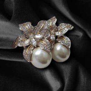 Puede incluir: Un par de pendientes con forma de flor. Cada pendiente presenta una gran perla blanca en forma de gota. El diseño floral está detallado con cristales transparentes y detalles dorados, sobre un fondo oscuro.
