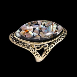 Könnte beinhalten: Ein goldener Ring mit einem großen ovalen Stein, der in einen schwarzen Hintergrund gefasst ist. Der Stein besteht aus irisierendem Perlmutt.