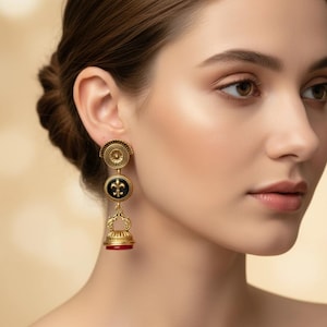 May include: Gold-toned drop earrings with a three-part design. The top section is a circular, detailed design. The middle section is a black circle with a gold fleur-de-lis. The bottom section is a bell shape with a red base.