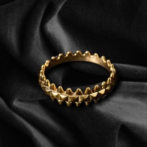 Könnte beinhalten: Ein goldfarbenes Armband mit einem sich wiederholenden Muster aus dreieckigen Spitzen. Das kreisförmige Armband liegt auf einer schwarzen, strukturierten Oberfläche. Das Schmuckstück ist ein Modeaccessoire.