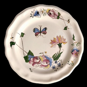 Pode incluir: Um prato de cerâmica branco com borda ondulada, decorado com motivos florais e borboletas pintados à mão. O design apresenta flores rosa, azuis e amarelas, e uma borboleta azul e rosa no centro. O prato tem uma estética vintage.