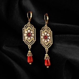 May include: A pair of gold-toned earrings with a vintage design. Each earring features a central red stone surrounded by small white pearls. The earrings have a rectangular shape with ornate detailing and a red teardrop-shaped bead at the bottom.