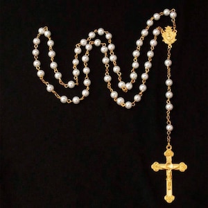 Peut inclure: Un chapelet de perles blanches avec une croix dorée et un pendentif doré. Le chapelet est disposé sur un fond noir.
