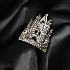 Peut inclure: Une bague argentée ornée d'un motif de cathédrale. La bague présente des éléments architecturaux détaillés, notamment des flèches, des arches et une rosace centrale. Elle est posée sur un fond noir texturé.