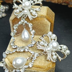 以下が含まれることがあります： 真珠とラインストーンで飾られた銀色のブローチ2つ。大きなブローチは、中央に大きな真珠が付いたボウのデザイン。小さなブローチは、中央に小さな真珠が付いた同様のボウのデザイン。両方のブローチには、ぶら下がっている真珠のドロップがあります。