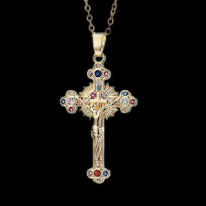 Puede incluir: Un collar con un colgante de cruz de tono dorado con un diseño detallado y piedras preciosas de colores. La cruz presenta una figura de Jesucristo y está adornada con piedras azules, rosas y rojas.