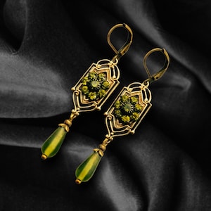 May include: A pair of gold-toned, Art Deco-style earrings. Each earring features a rectangular frame with green gemstone accents and a teardrop-shaped, green and yellow pendant. The earrings are displayed against a black, textured background.