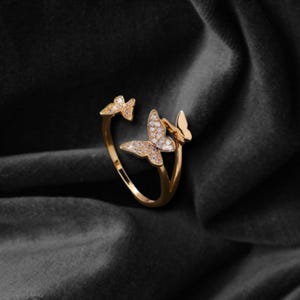 May include: A gold-colored ring featuring three butterfly designs. The central butterfly is adorned with small, sparkling stones. The ring is set against a dark, textured background, highlighting its intricate details and elegant design.