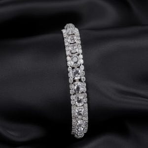 Può includere: Un bracciale d'argento con numerose pietre chiare e scintillanti di varie forme e dimensioni. Il bracciale è posto su uno sfondo nero e setoso, che ne evidenzia la brillantezza e il design. Il bracciale è un gioiello.