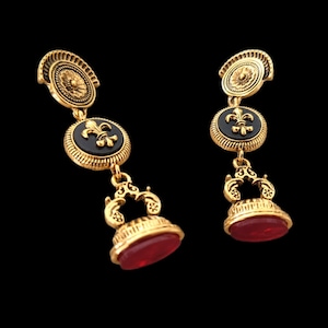 Peut inclure: Boucles d'oreilles pendantes dorées avec des accents noirs et rouges. Les boucles d'oreilles présentent un motif de fleur de lys et une pierre précieuse rouge.