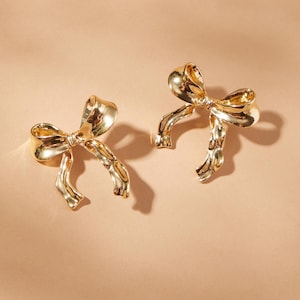 May include: A pair of gold-toned bow-shaped earrings. The earrings are made of metal and have a textured finish.