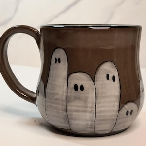 May include: A brown ceramic mug featuring a hand-painted design of multiple white ghost figures with simple black dot eyes. The ghosts are arranged in a group around the exterior of the mug, which has a smooth, glossy finish and a curved handle.