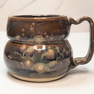 May include: A handcrafted ceramic mug with a unique stacked design. The mug features a rich, dark brown glaze with hints of purple and green, creating a textured, organic appearance. The handle is curved and integrated into the design.
