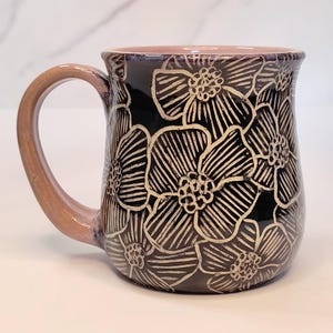 May include: A ceramic mug with a glossy black glaze featuring a carved, cream-colored floral pattern across the exterior. The mug has a curved handle and an interior rim finished in a soft, muted pink tone, providing a contrast to the dark body.