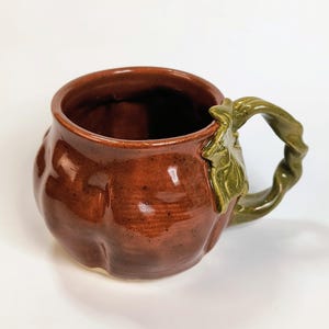 May include: A glossy, round, orange-brown ceramic mug shaped like a pumpkin with vertical ridges. The mug features a sculpted, twisted green handle that resembles a vine, complete with a small, textured green leaf detail attached at the base.