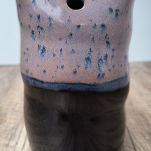 May include: A ceramic vase with a speckled pink and blue glaze on the top half and a solid black glaze on the bottom half. The vase has a round opening at the top.