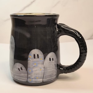 May include: A black ceramic mug featuring a glossy finish and a textured handle. The exterior is decorated with hand-painted light gray ghost figures, one of which displays a pattern of small blue flowers and yellow dots against the dark background.
