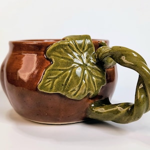 May include: A glossy brown ceramic mug with a rounded body, featuring a sculpted green leaf attached to the side and a handle shaped like a twisted green vine. The surface of the mug has a smooth, reflective finish with subtle dark speckles throughout.