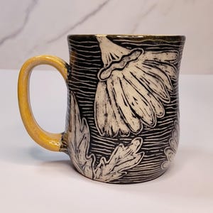 May include: Ceramic mug with a yellow handle and a black and white floral design. The mug features a large white flower and leaves against a black background with horizontal lines. The handle is a solid yellow color.