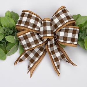 May include: A decorative bow with a brown and white checkered pattern, set against a backdrop of green foliage. The bow has a burlap-textured trim and is centered on a white surface, creating a rustic aesthetic.