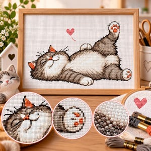 Könnte beinhalten: Eine Kreuzstichstickerei einer grau-weißen Katze, die auf dem Rücken liegt, mit einem kleinen rosa Herz darüber, gerahmt in hellem Holz. Das Design zeigt detaillierte Stiche des Katzengesichts, Pfoten mit orangefarbenen Ballen und Fell.