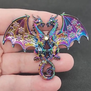 May include: A colorful dragon brooch with iridescent wings and a swirling tail. The dragon's body is a mix of blues, greens, and purples, with a central jewel. The wings are detailed with floral patterns. This fantasy-themed accessory is a unique piece of jewelry.