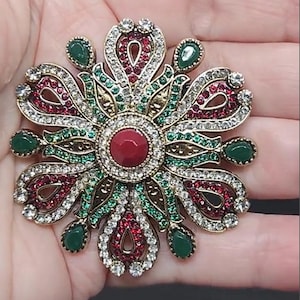 May include: Ornate, gold-toned brooch in a floral design. The brooch features a central red stone surrounded by a ring of clear crystals. The petals are adorned with red and green stones, and clear crystals. Green teardrop-shaped stones are placed around the perimeter.