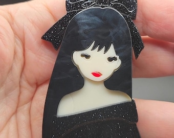 Broche acrílico para mujer con cabello negro, blusa negra brillante y lazo para el cabello.