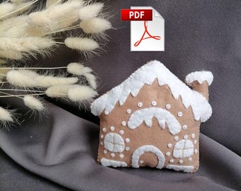 Pode incluir: Um enfeite de casa de gengibre de feltro com detalhes de glacê branco e uma chaminé. O enfeite está sobre um fundo de tecido cinza. O texto PDF é visível no canto superior direito da imagem.
