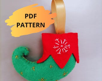 May include: A felt Christmas elf shoe ornament in green and red, with gold star accents and a white snowflake design. The ornament has a gold ribbon for hanging and the text "PDF PATTERN" in a yellow and orange shape.