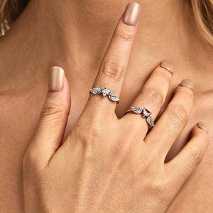 Op de afbeelding: Twee zilveren ringen met heldere en roze stenen. De ring op de wijsvinger heeft een ontwerp van drie verbonden bladeren met heldere stenen. De ring op de middelvinger heeft een ontwerp van twee verbonden vleugels met een roze steen in het midden.