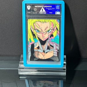 May include: A graded drawing of a female anime character with green hair and blue eyes. The drawing is in a blue and white graded card with the text "WIZSTUDIOS" and "EVERYONE" on it.