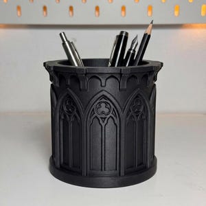 May include: A matte black cylindrical pencil holder featuring a relief pattern of pointed gothic arches and trefoil motifs. The container holds four writing instruments, including a silver metal pen, two black pens, and a wooden graphite pencil.