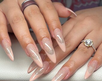 Peut inclure: Gros plan sur une main avec de longs ongles en amande. Les ongles sont peints d'une couleur rose pâle avec un recouvrement de paillettes transparentes. Les ongles sont accentués par une bague argentée à l'index et une bague en diamant à l'annulaire.